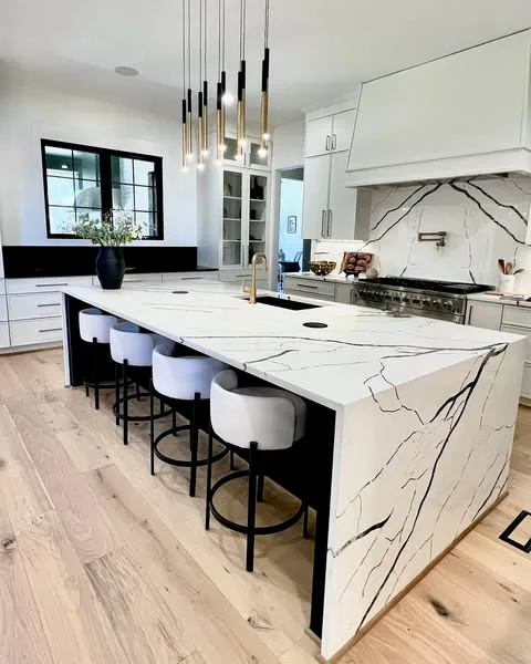 White kitchen with a veined waterfall island, bar seating and black pendant lights.